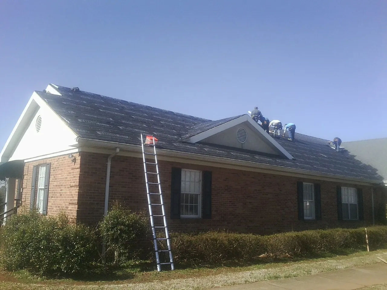 Crew tearing shingles off a brick ranch roof with a ladder set — Emergency Roof Repair work by our crew in Commack