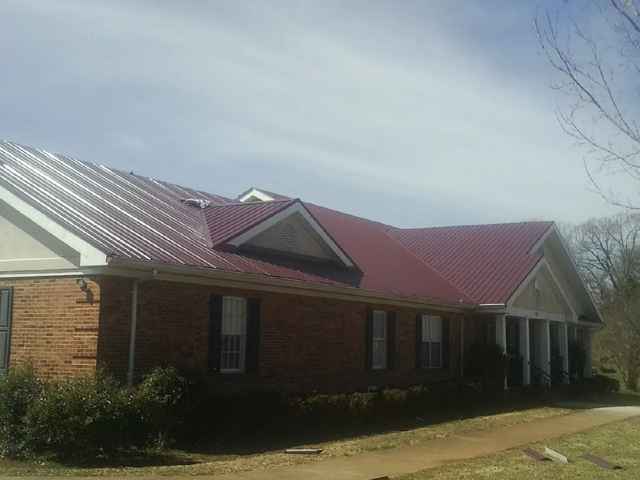 Red metal roof with gabled entries on a long brick building, from a Hail Damage Roof Repair job in Commack