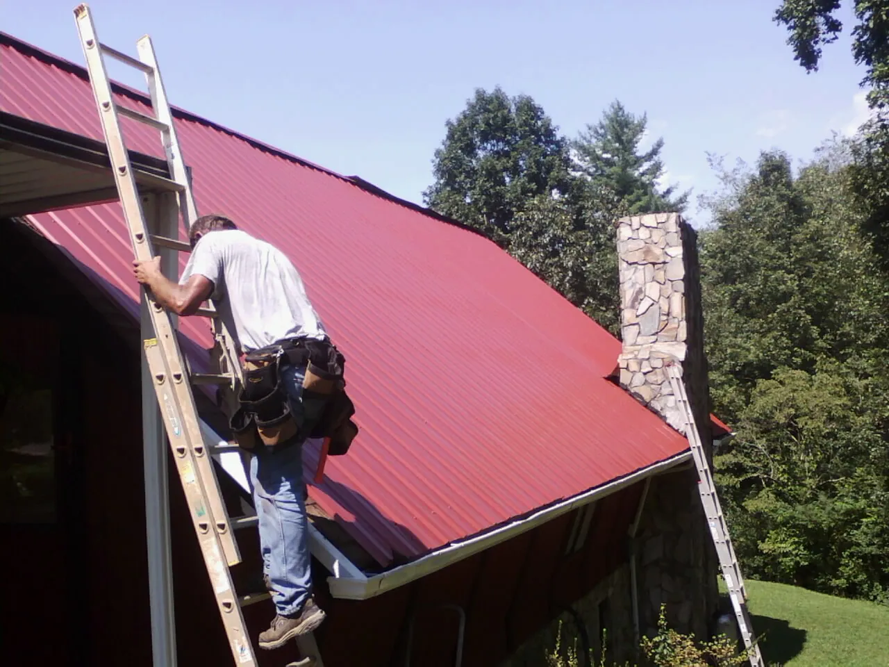 Roofer on a ladder beside a red metal roof and stone chimney — Soffit & Fascia Repair by Roofing Brothers in Commack
